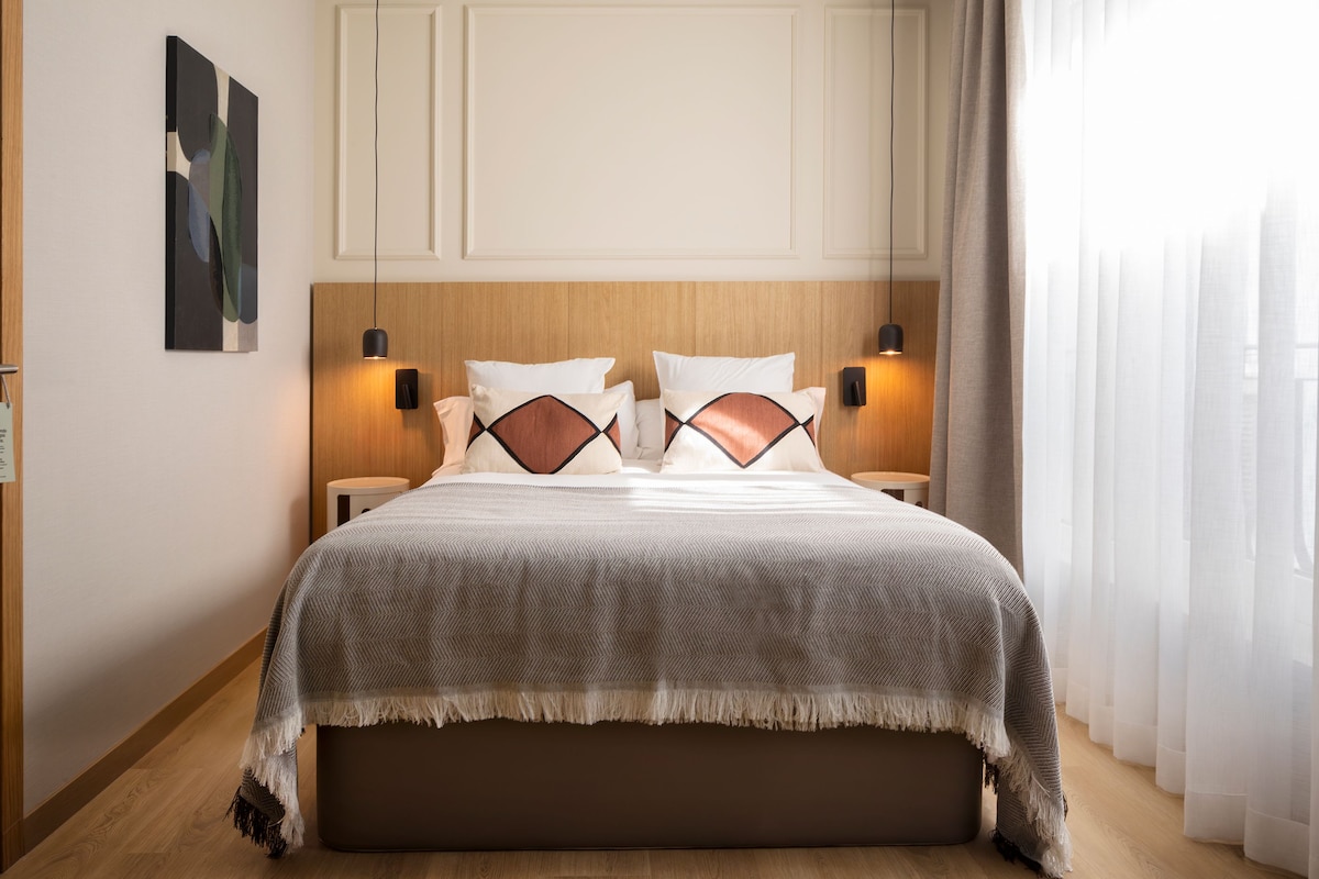 Un dormitorio con suelo de madera, una cama con colcha estampada, dos lámparas de noche y un cuadro en la pared.