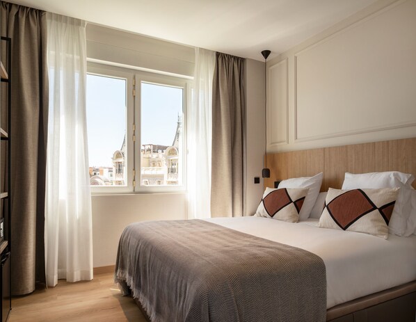 Habitación de hotel con una cama grande, una ventana con cortinas, un cabecero de madera y vistas a edificios.