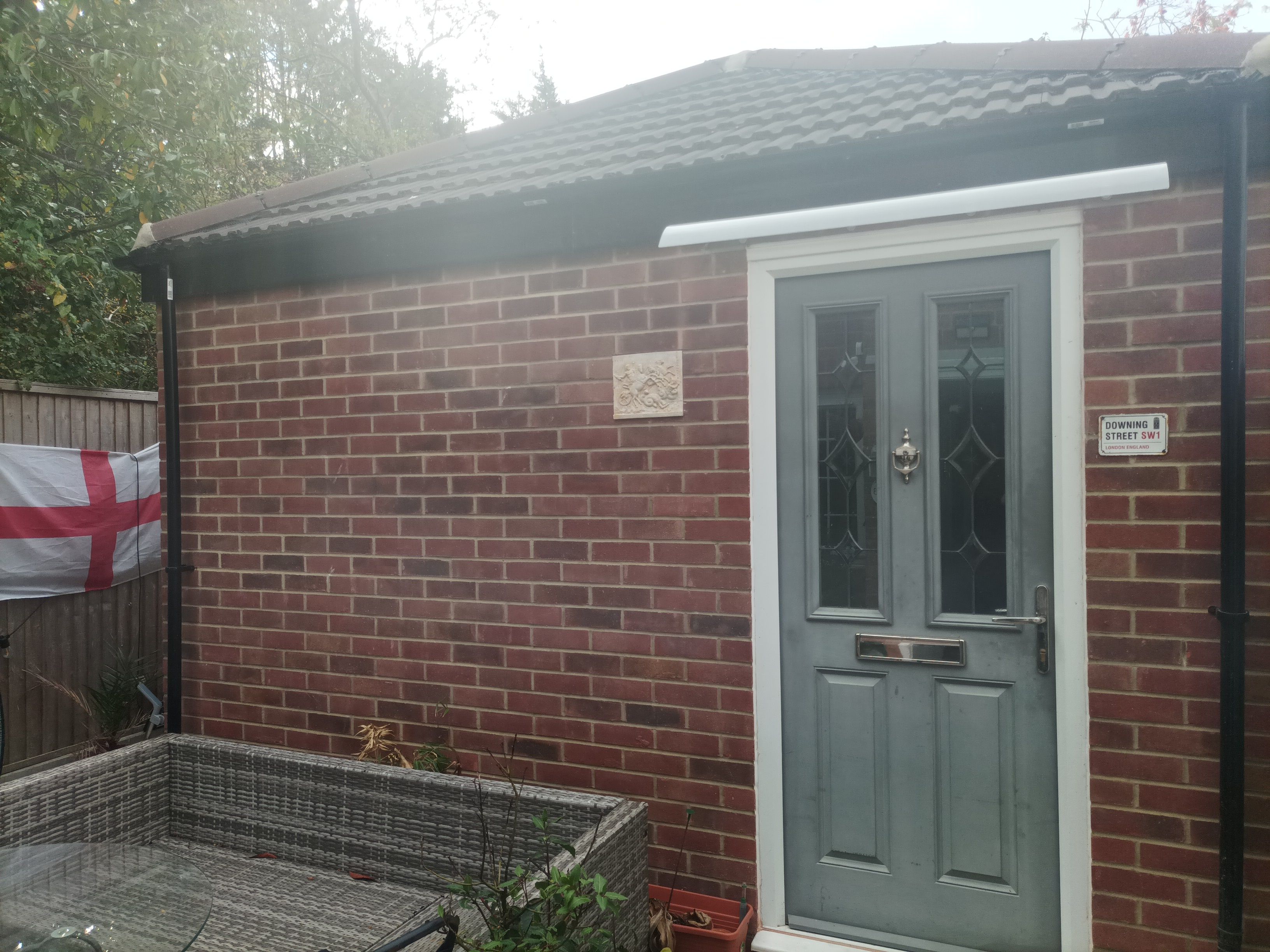 A brick building with a grey door, a small window, and a sign indicating 'Downing Street SW1 London, England'.