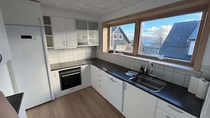 A kitchen with white cabinets, a black countertop, a built-in oven, a sink, and a window with a view of houses. - Tórshavn Central Guesthouse (Tórshavn)