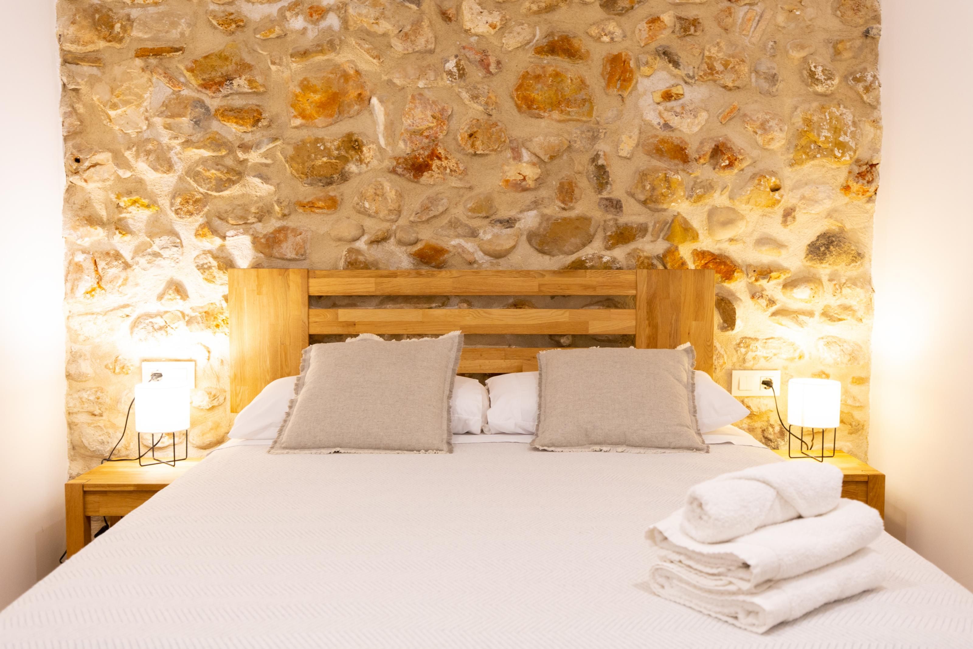 A bedroom with a stone wall, wooden headboard, white bedding, and two bedside lamps.