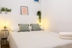 Un lit bien fait, avec deux oreillers, une lampe de chevet, une petite table ronde et une étagère murale agrémentée de plantes.