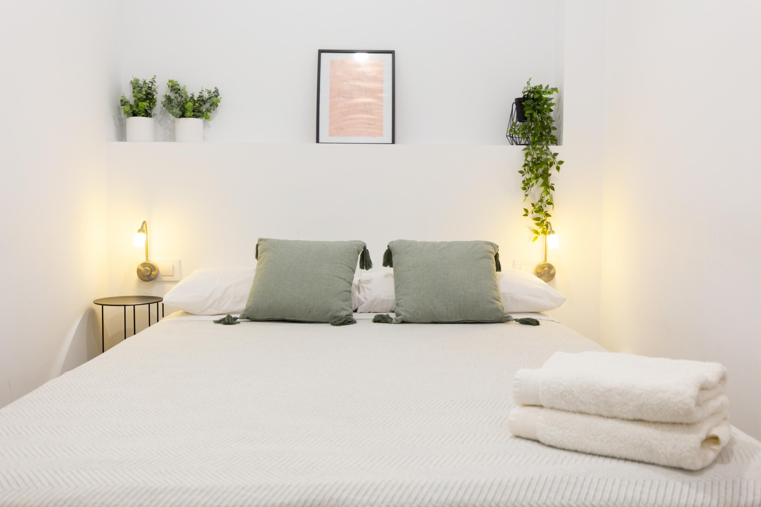 A hotel room with a bed, two bedside lamps, a framed picture on the wall, and two potted plants.