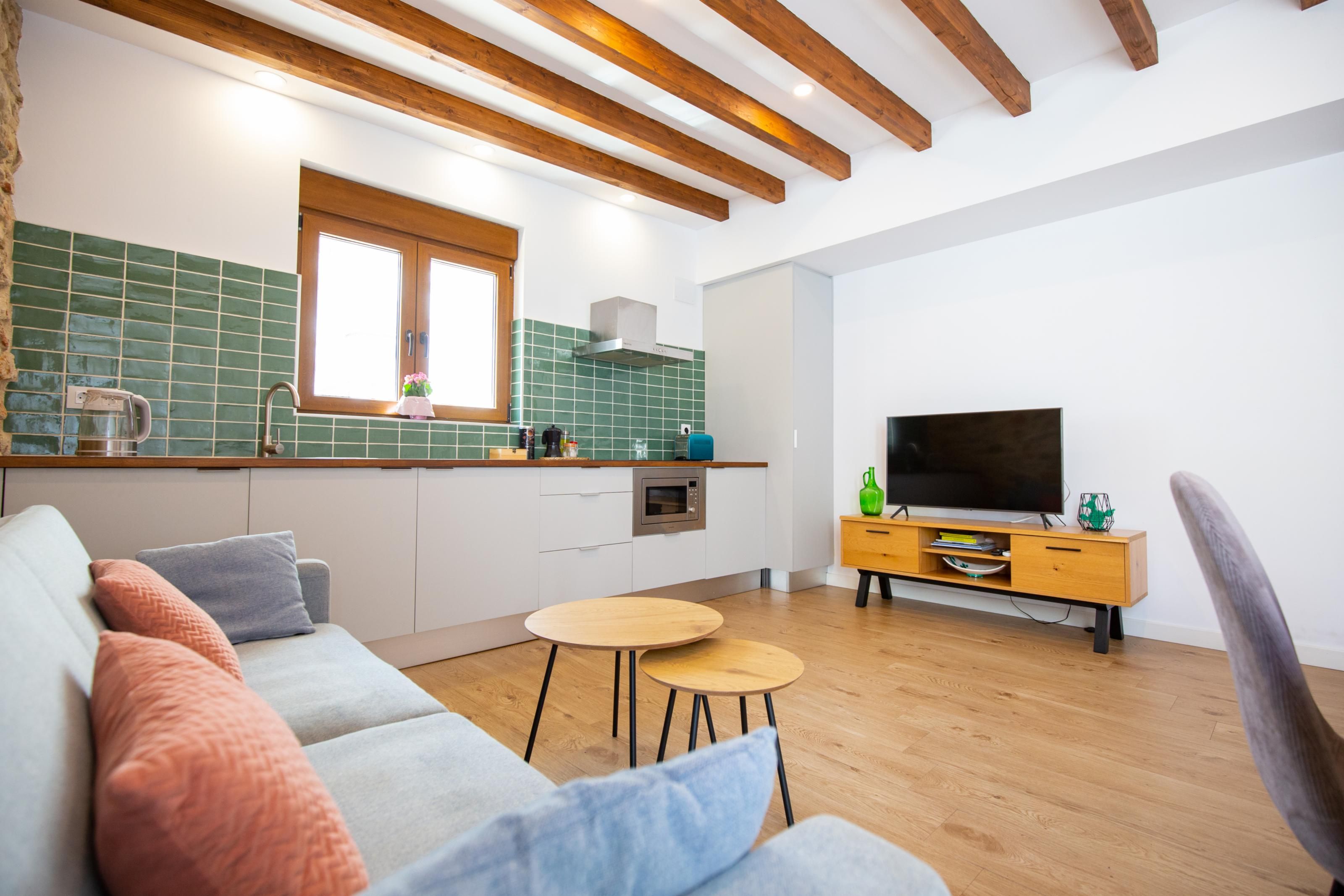 Une cuisine moderne avec une fenêtre, des poutres en bois au plafond, un four encastré et un téléviseur posé sur un meuble en bois.