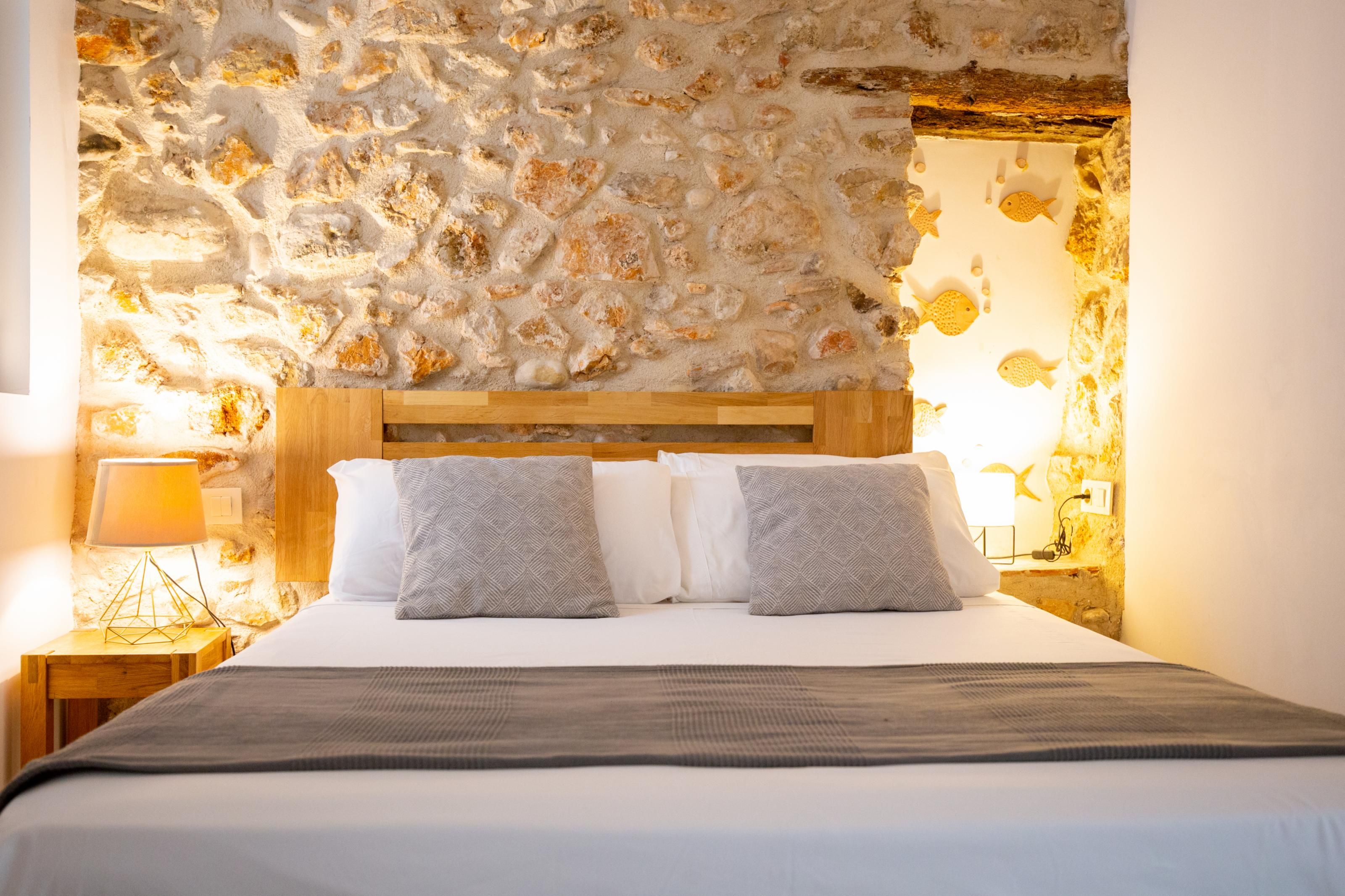 Una cama con cabecera de madera, dos almohadas y una colcha en una habitación con pared de piedra texturizada.