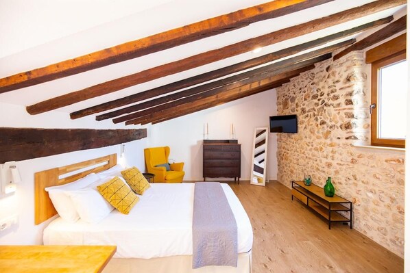 Un dormitorio con techo inclinado, una cama con ropa de cama blanca, una silla amarilla, una mesita de noche de madera y una pared de piedra.