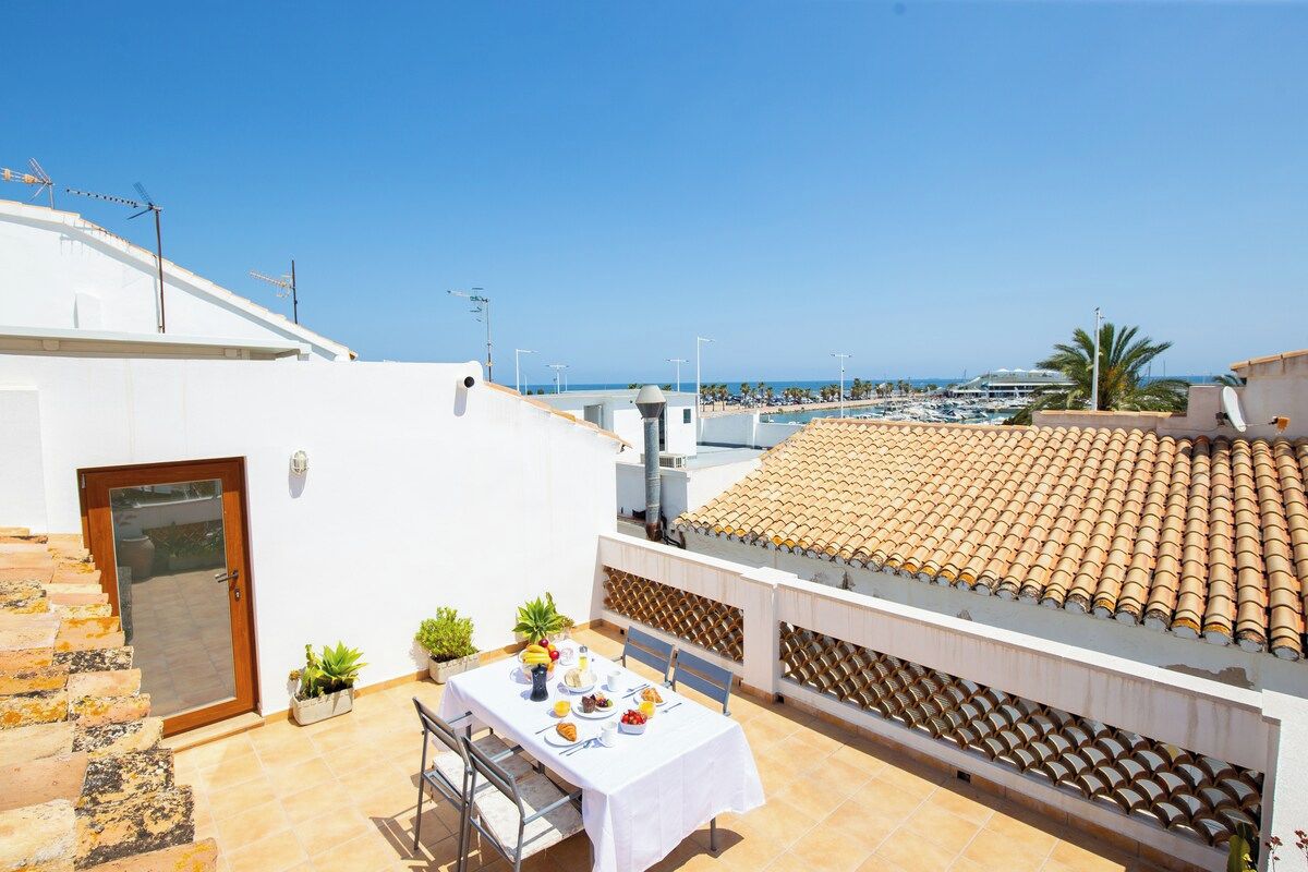 Terraza en la azotea con una mesa lista para comer, con vistas a un puerto deportivo y al mar.