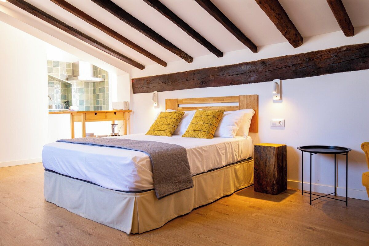 Un dormitorio con cama, vigas de madera, una mesita de noche y un ventanal con vistas.