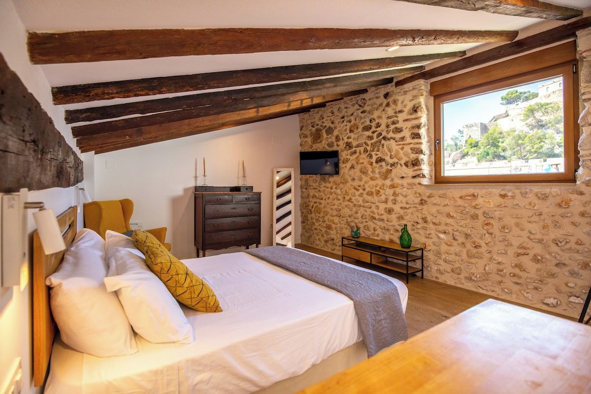 Un dormitorio con pared de piedra, vigas de madera, cama con almohadas, cómoda, televisor y ventana con vistas.