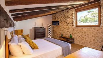 Un dormitorio con pared de piedra, vigas de madera, cama con almohadas, cómoda, televisor y ventana con vistas.