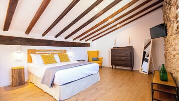 Un dormitorio con techo de madera, una cama con ropa blanca, una silla amarilla, un aparador y un televisor en la pared.