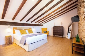 Un dormitorio con techo de madera, una cama con ropa blanca, una silla amarilla, un aparador y un televisor en la pared.