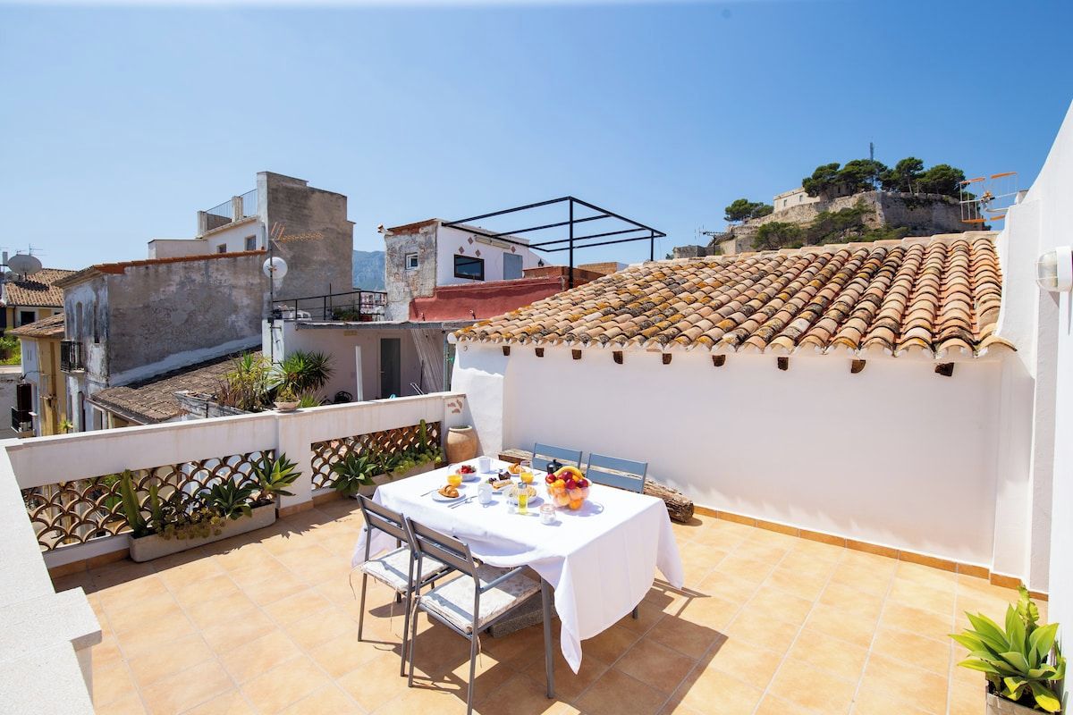 Una terraza en la azotea con una mesa dispuesta para comer, rodeada de plantas en macetas y vistas a un paisaje urbano.
