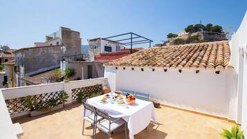 Una terraza en la azotea con una mesa dispuesta para comer, rodeada de plantas en macetas y vistas a un paisaje urbano.