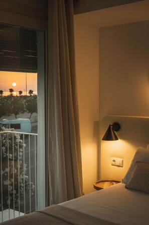 Quarto de hotel com cama, mesa de cabeceira, luminária e vista do pôr do sol pela janela.