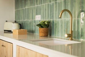 Cozinha com bancada branca, torneira dourada, painel de azulejos verdes, torradeira e uma pequena planta em cesto.
