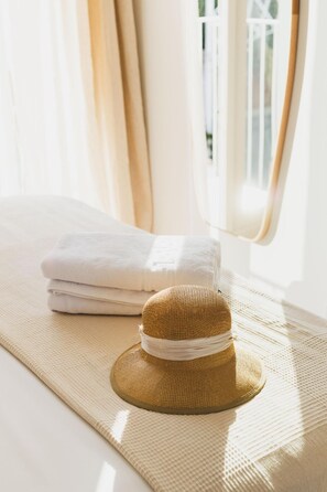 Quarto com janela, toalha branca enrolada e chapéu de palha sobre um tapete tecido.