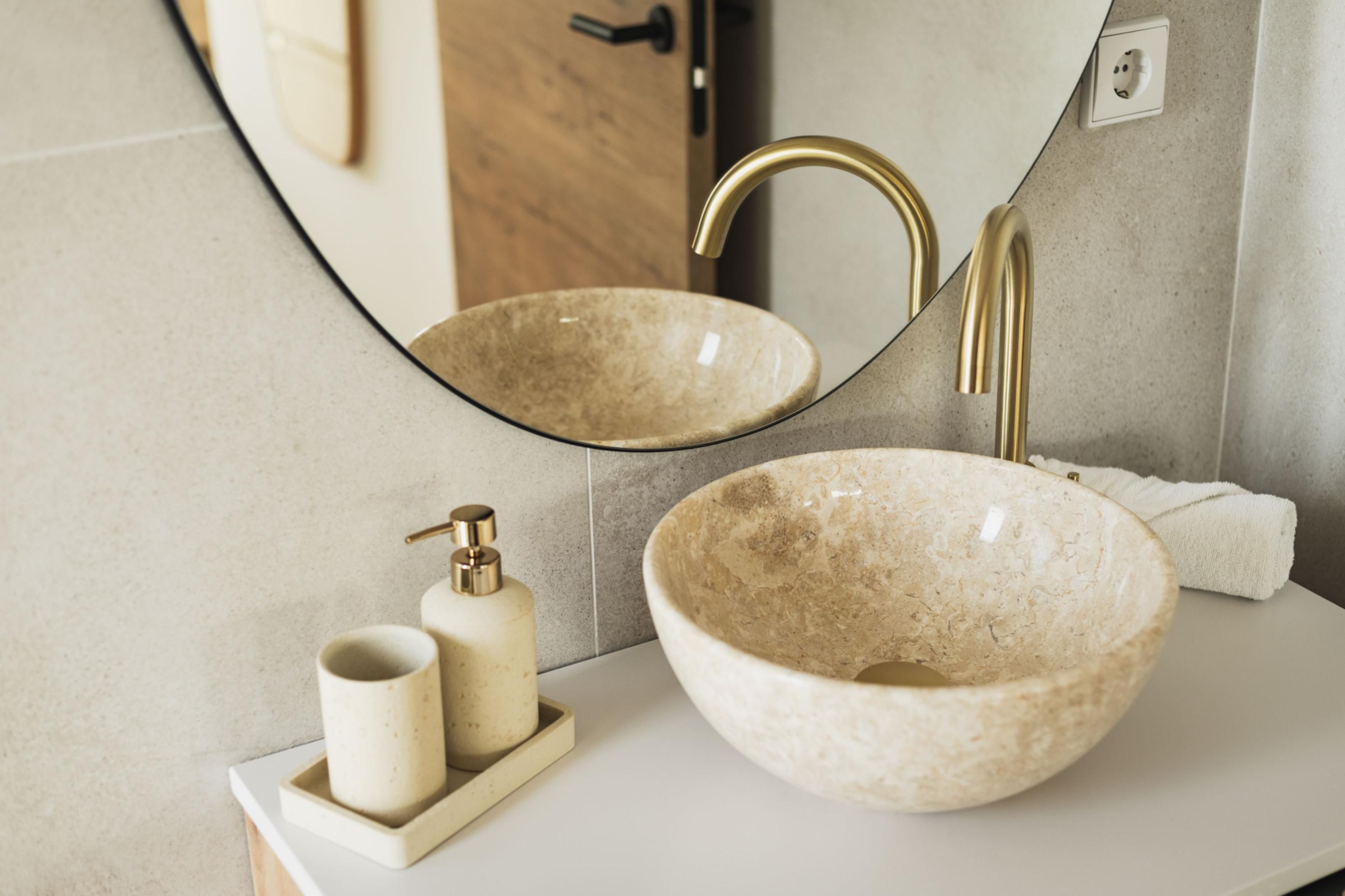 Un baño con un lavabo beige redondeado, un grifo dorado y un espejo que refleja el lavabo. Hay un dispensador de jabón y una toalla en la encimera.