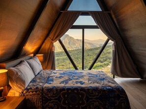 Quarto com cama, almofadas, abajur e vista para montanhas pela janela.