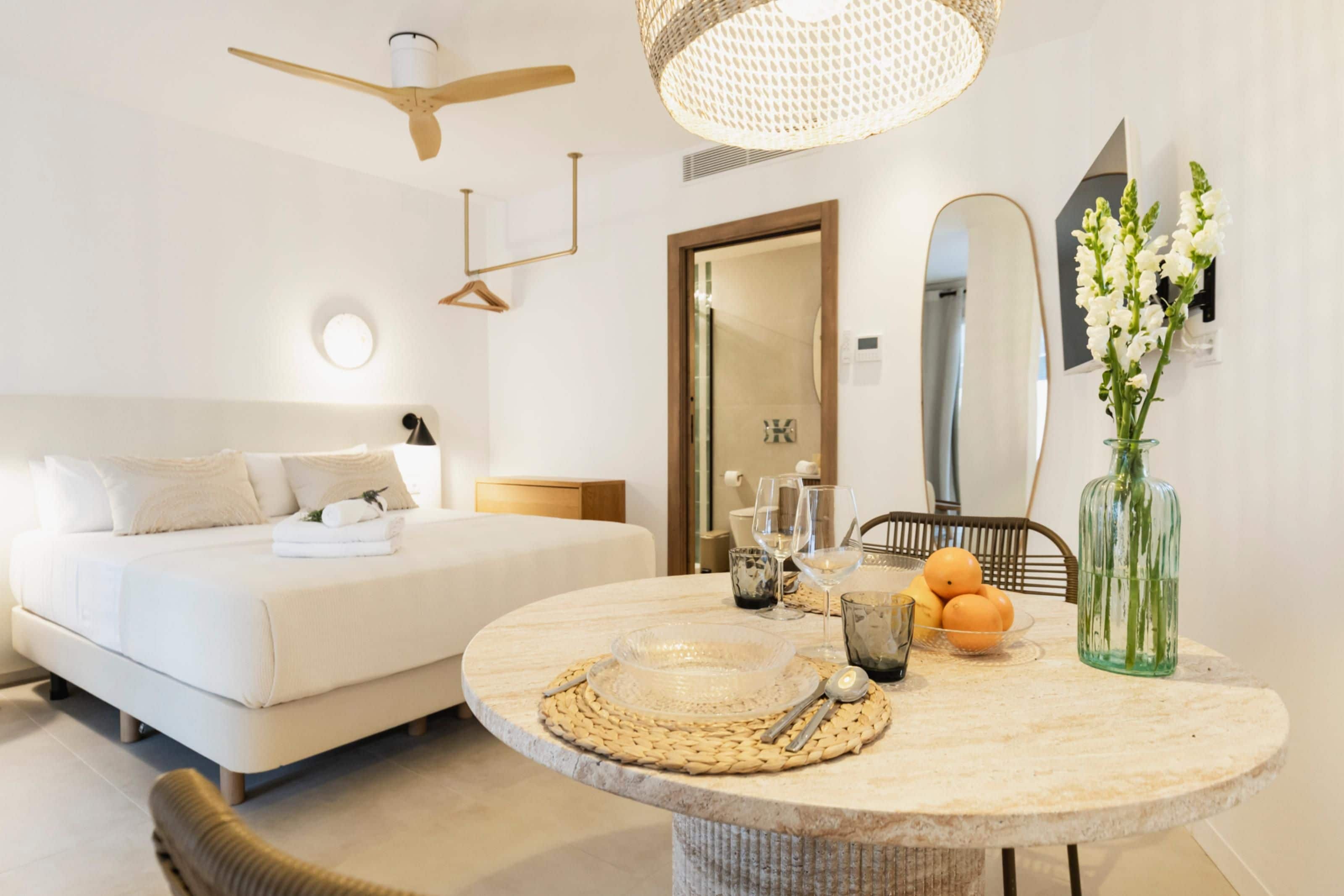 Een moderne hotelkamer met een bed, een ronde eettafel met placemat, een vaas met bloemen en een spiegel aan de muur.