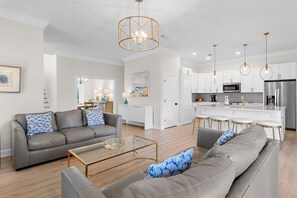 Een moderne woonkamer met een grijze bank, een salontafel en een keukenhoek met witte kasten en roestvrijstalen apparatuur.