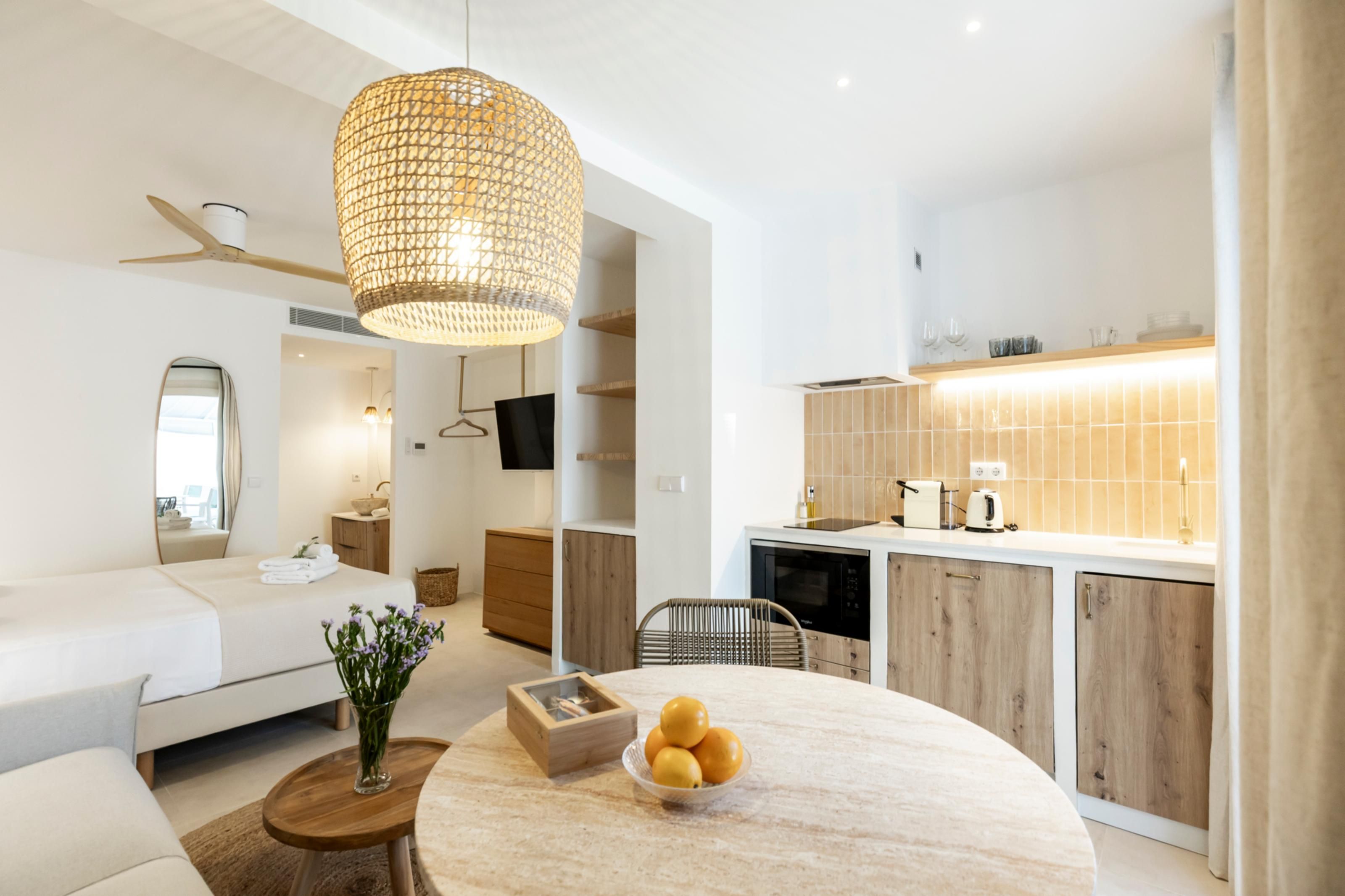 A modern hotel room with a sofa, a dining table with fruit, a hanging light fixture, and a kitchen area with a microwave and toaster.