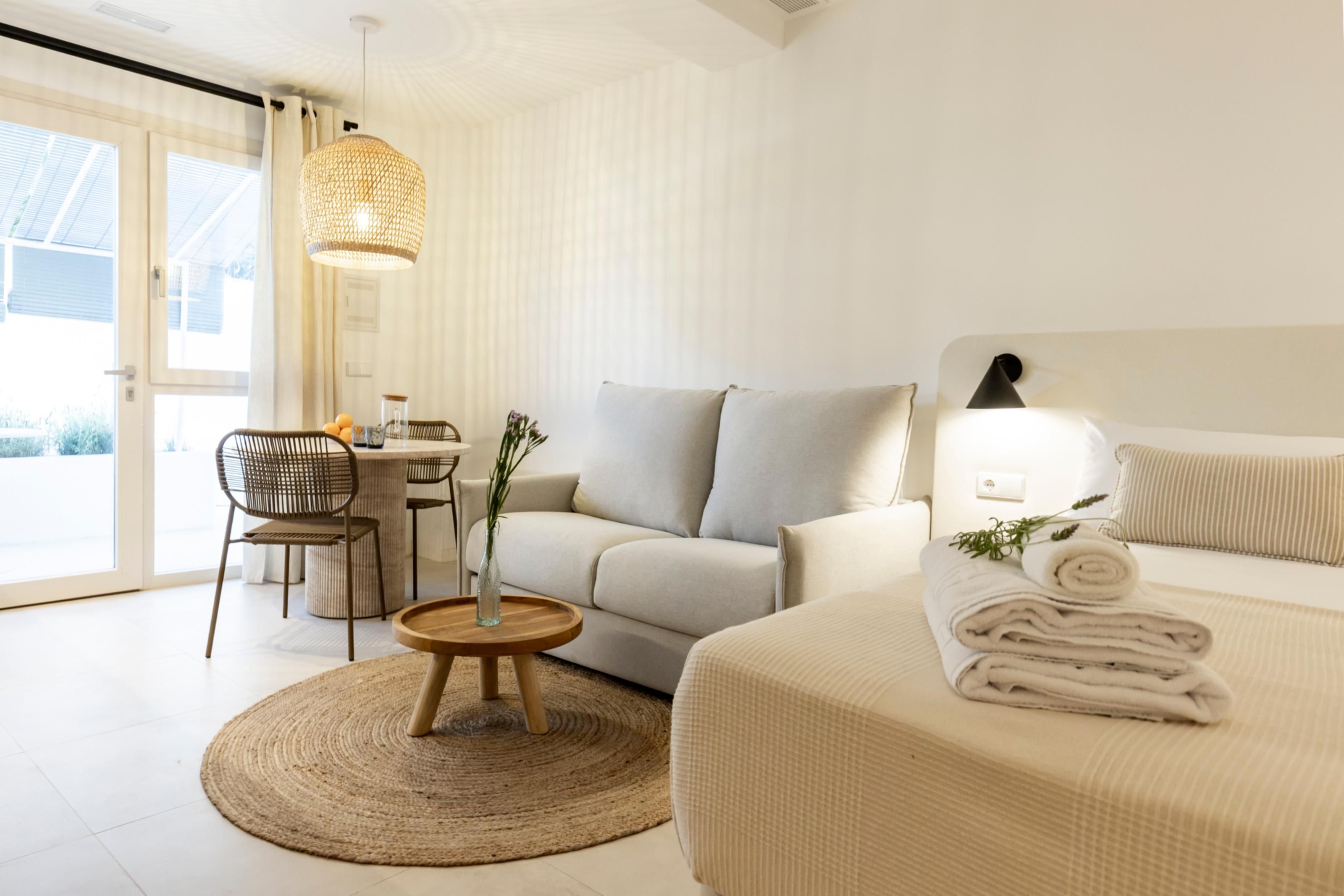A modern hotel room with a sofa, a dining table, a chair, a small round table with a vase, and a bed with rolled towels.