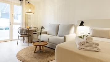 A modern hotel room with a sofa, a dining table, a chair, a small round table with a vase, and a bed with rolled towels.