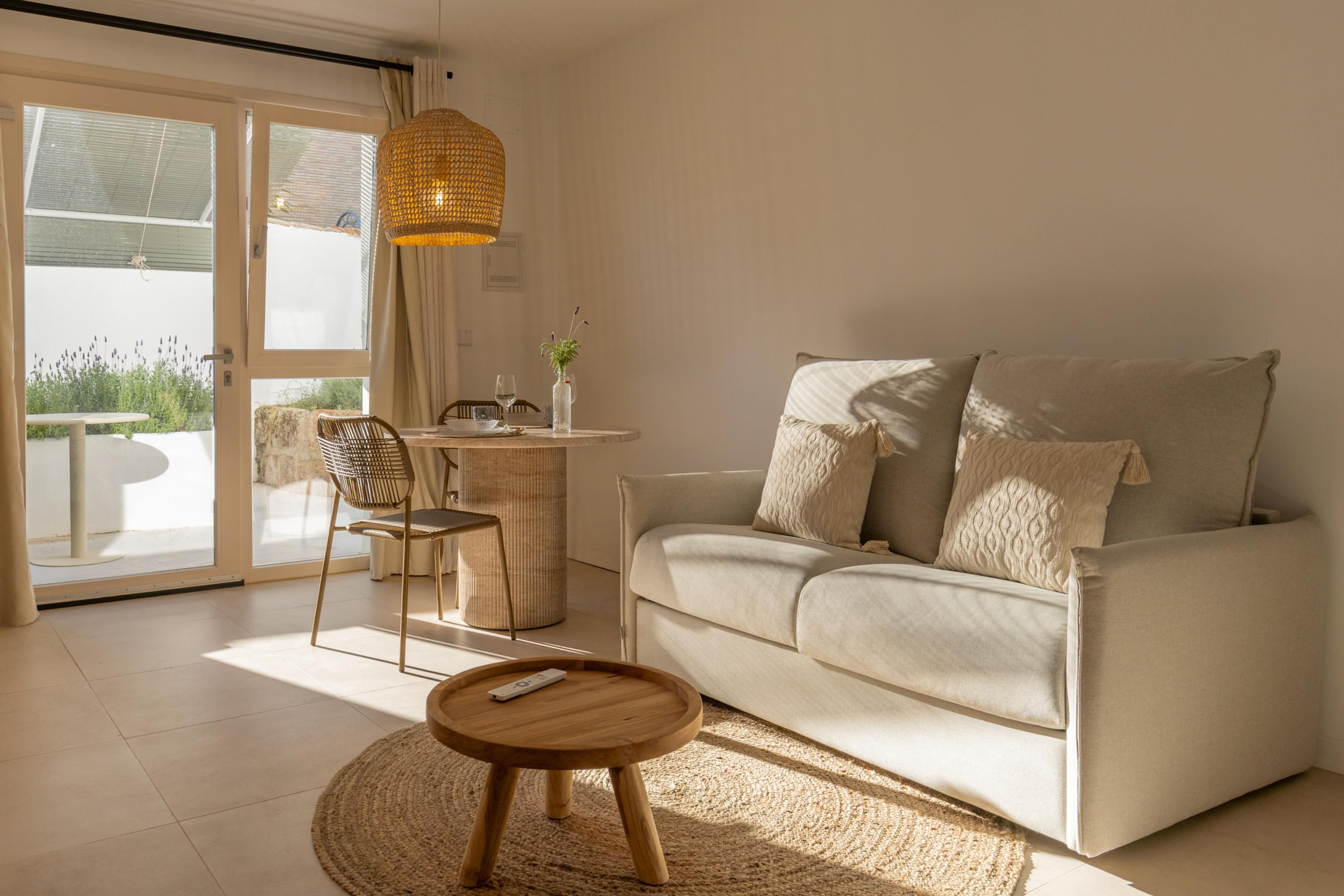 A living room with a sofa, a round wooden table, a wicker chair, a hanging light fixture, and a view through a glass door.