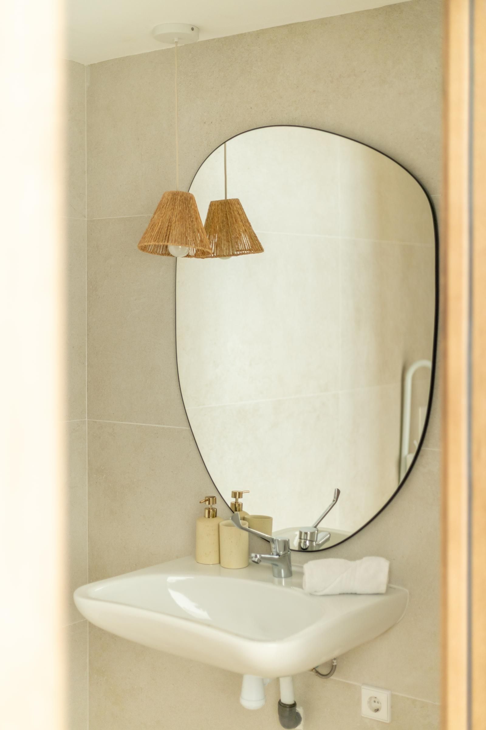Un baño con un espejo ovalado grande, un lavamanos blanco, dos dispensadores dorados y una lámpara colgante.