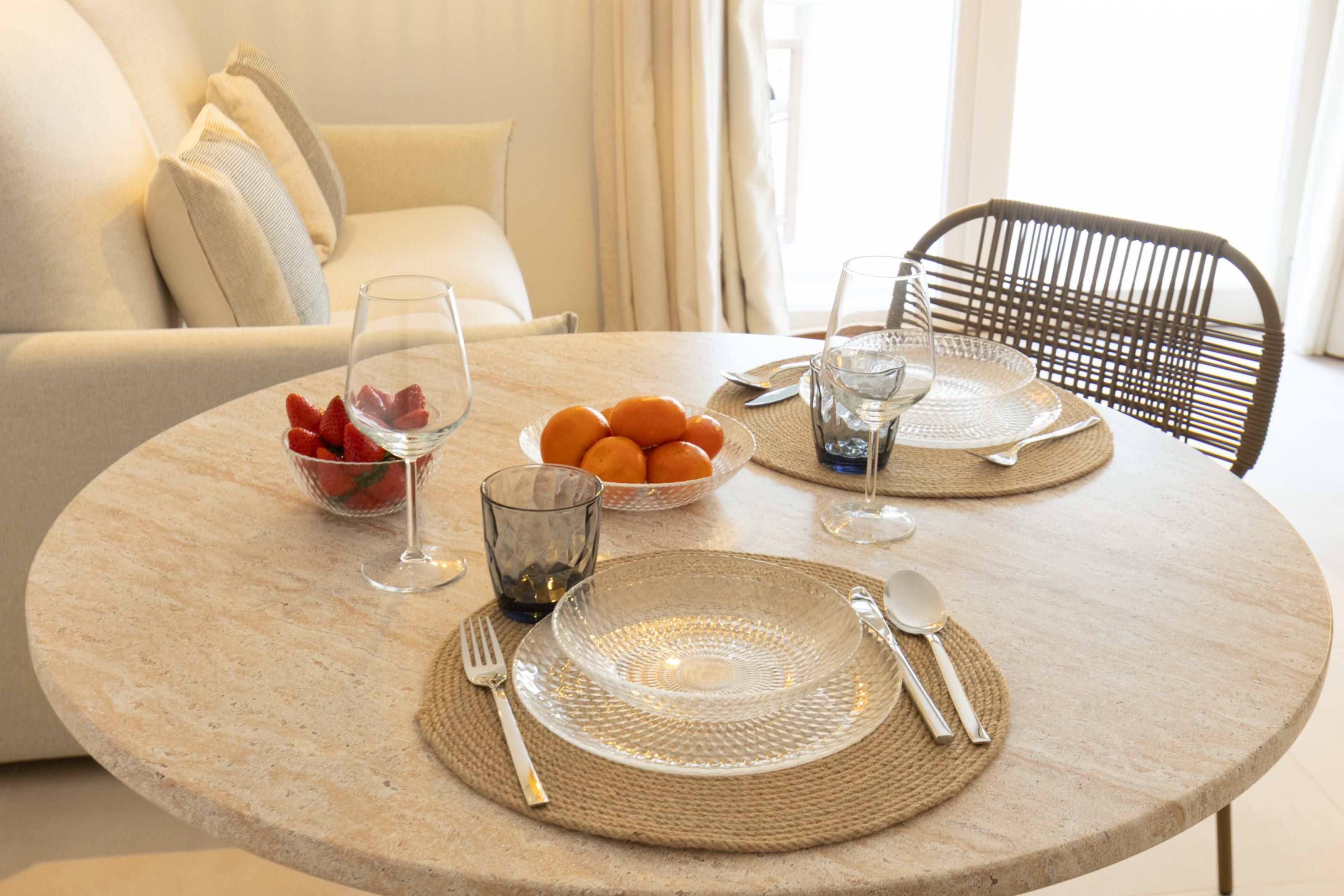 Een ronde eettafel met een geweven placemat, een schaal aardbeien, een schaal mandarijnen en een glas water.