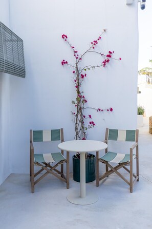 Uma pequena área externa com assento, incluindo uma mesa redonda, duas cadeiras e um vaso com flores rosa, além de uma parede branca.