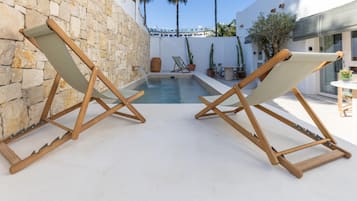 Twee houten lounge stoelen met canvas zittingen gericht op een zwembad, tegen een stenen muur en een wit gebouw met blauw dak.