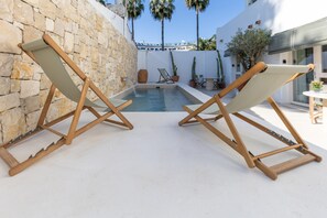 Duas poltronas de madeira com assentos de lona voltadas para uma piscina, em frente a um muro de pedra e um edifício branco com telhado azul.