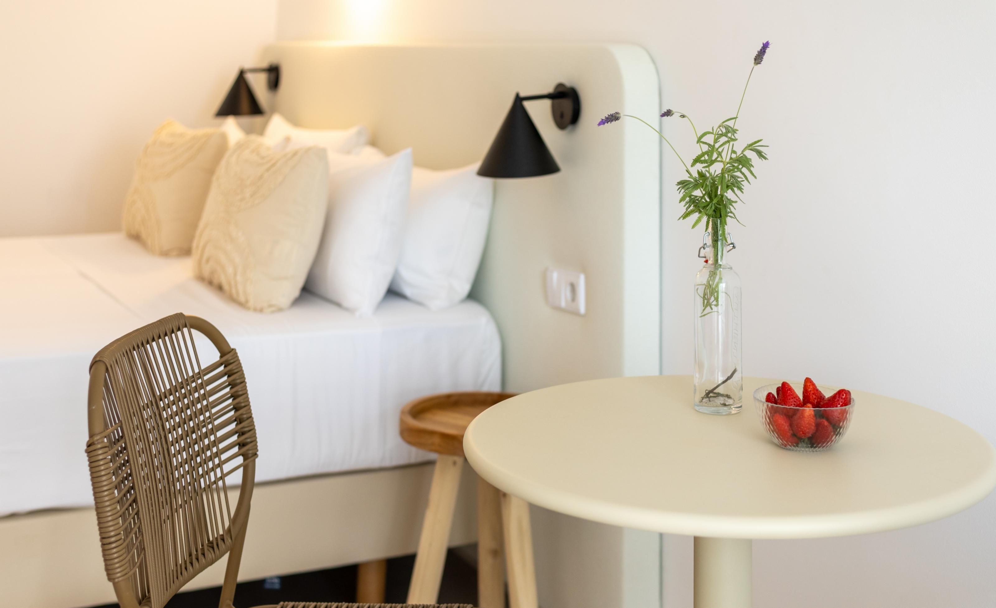 Een slaapkamer met een bed, een stoel, een tafel met een vaas en aardbeien, en een wandlamp.