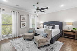 Une chambre à coucher comprenant un lit, deux fauteuils, une table de chevet, un ventilateur de plafond et une fenêtre avec des stores.