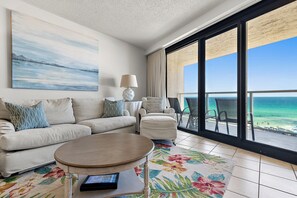 Ein Zimmer mit einer Couch, einem Sessel und einem Tisch mit einem Buch. An der Wand hängt ein Gemälde und durch große Fenster ist der Ozean zu sehen.