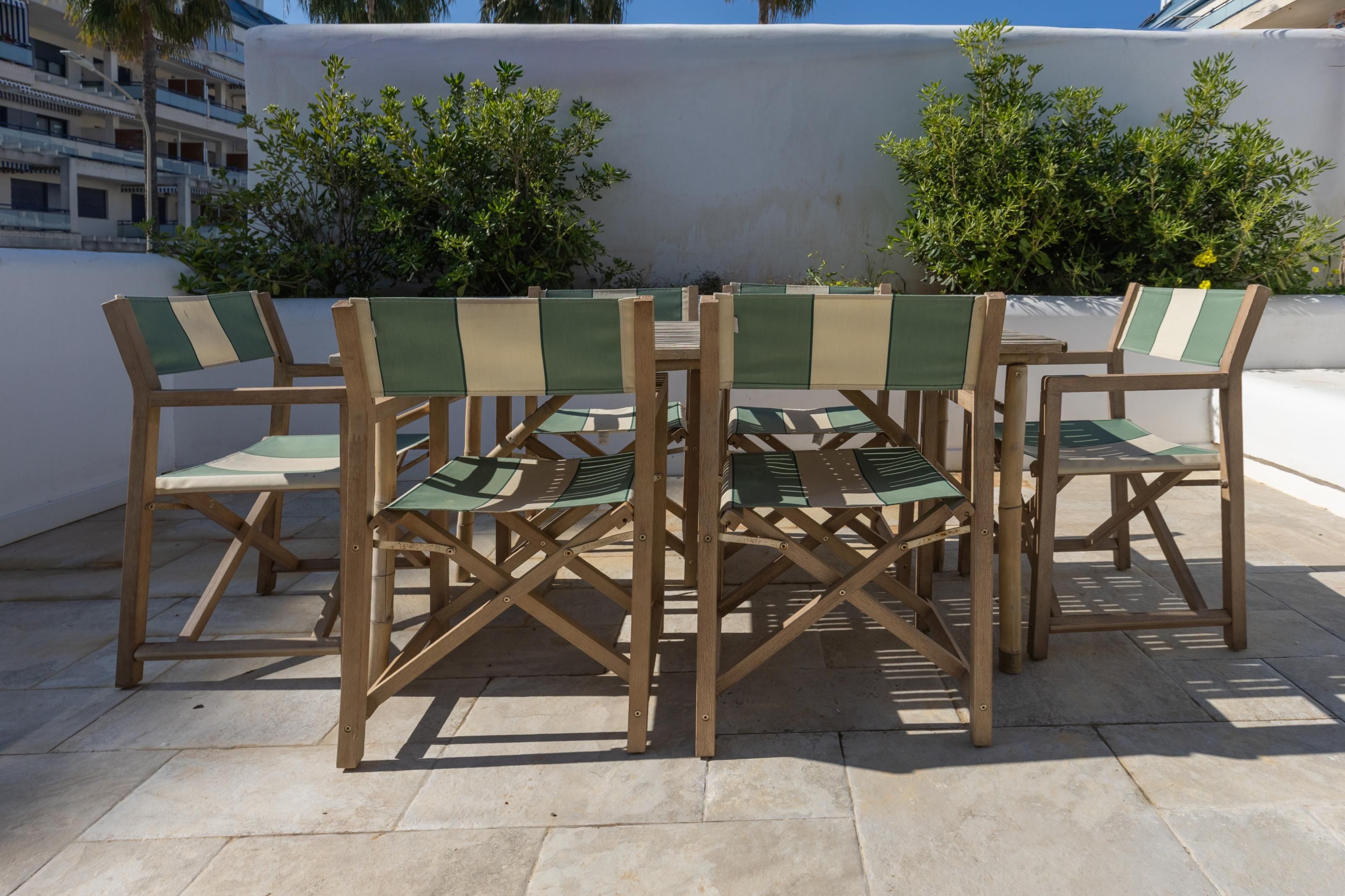 Zes houten stoelen met groene en beige kussens staan rond een kleine ronde tafel op een betegeld terras.