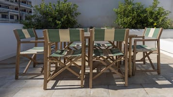 Zes houten stoelen met groene en beige kussens staan rond een kleine ronde tafel op een betegeld terras.