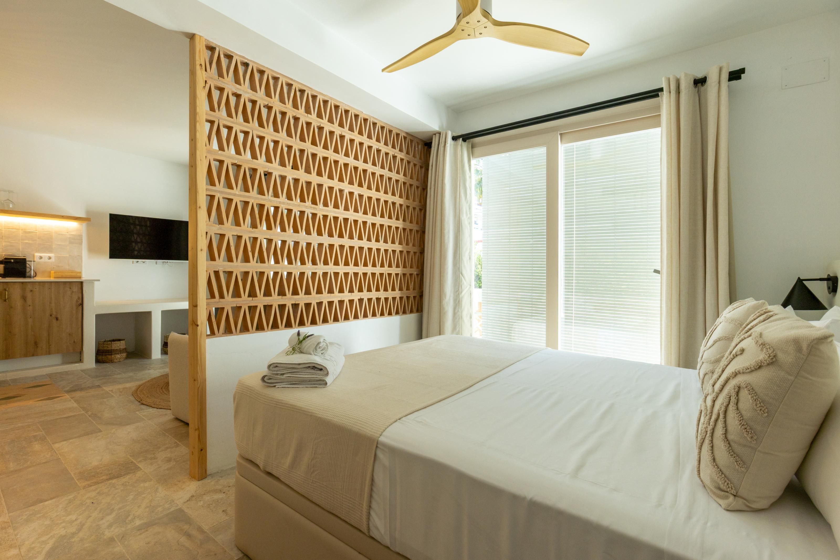 Een slaapkamer met een bed, een houten roomdivider, een bureau met een flatscreen televisie en een raam met gordijnen.