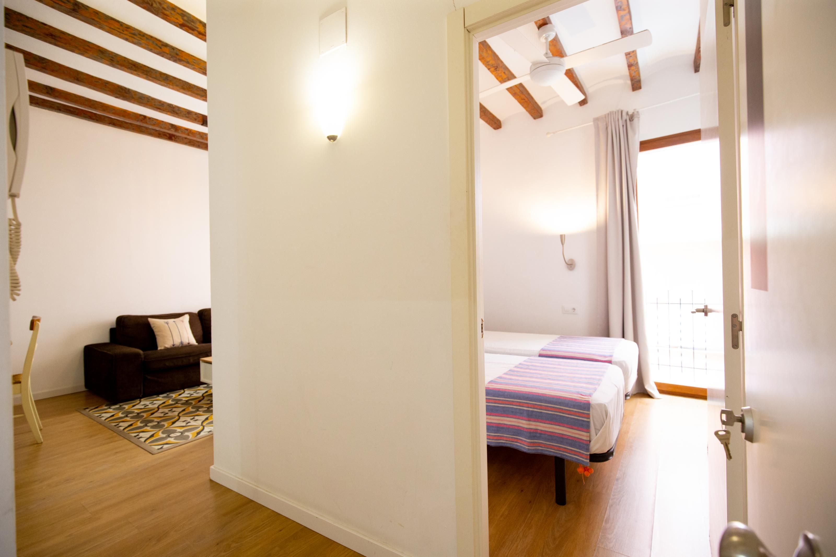 A room with a wooden floor, a sofa, a patterned rug, a bed with striped bedding, and a door leading to another room.