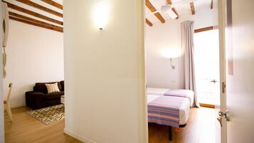 A room with a wooden floor, a sofa, a patterned rug, a bed with striped bedding, and a door leading to another room.