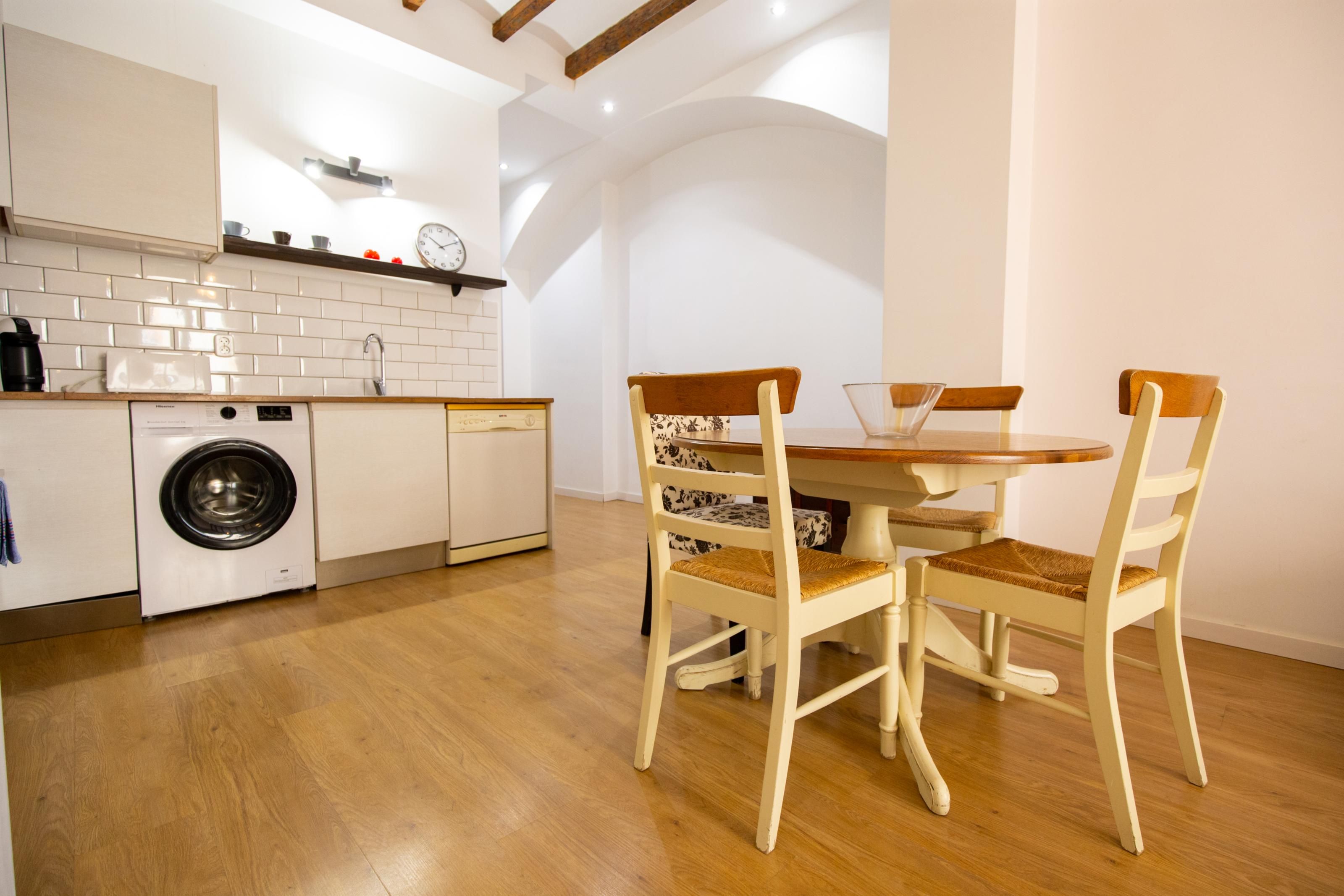 A compact kitchen with a washing machine, a dining table with chairs, and a clock on the wall.