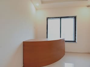 A room with a curved wooden desk, a window with blinds, and a plain white wall. - Collection O White House Regency (Nagpur)