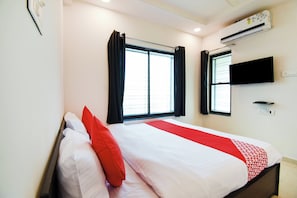Habitación de hotel con cama, dos ventanas con cortinas, unidad de aire acondicionado, televisor de pantalla plana montado en la pared y un enchufe.