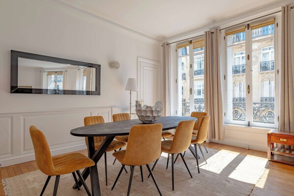 A dining area with a round table, six chairs, a mirror, and a view of a building through large windows. - Prestige Trémoille | Luxury 3BDR in the 8th Arron. (Paris)