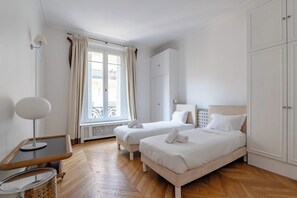 A hotel room with two beds, a desk, a chair, and a wardrobe. - Prestige Trémoille | Luxury 3BDR in the 8th Arron. (Paris)