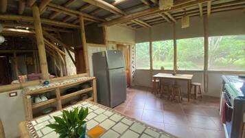 A kitchen with a bamboo ceiling, a refrigerator, a dining table with stools, and a view of greenery outside.