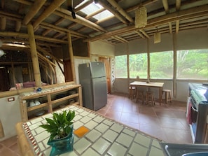 A kitchen with a bamboo ceiling, a refrigerator, a dining table with stools, and a view of greenery outside.
