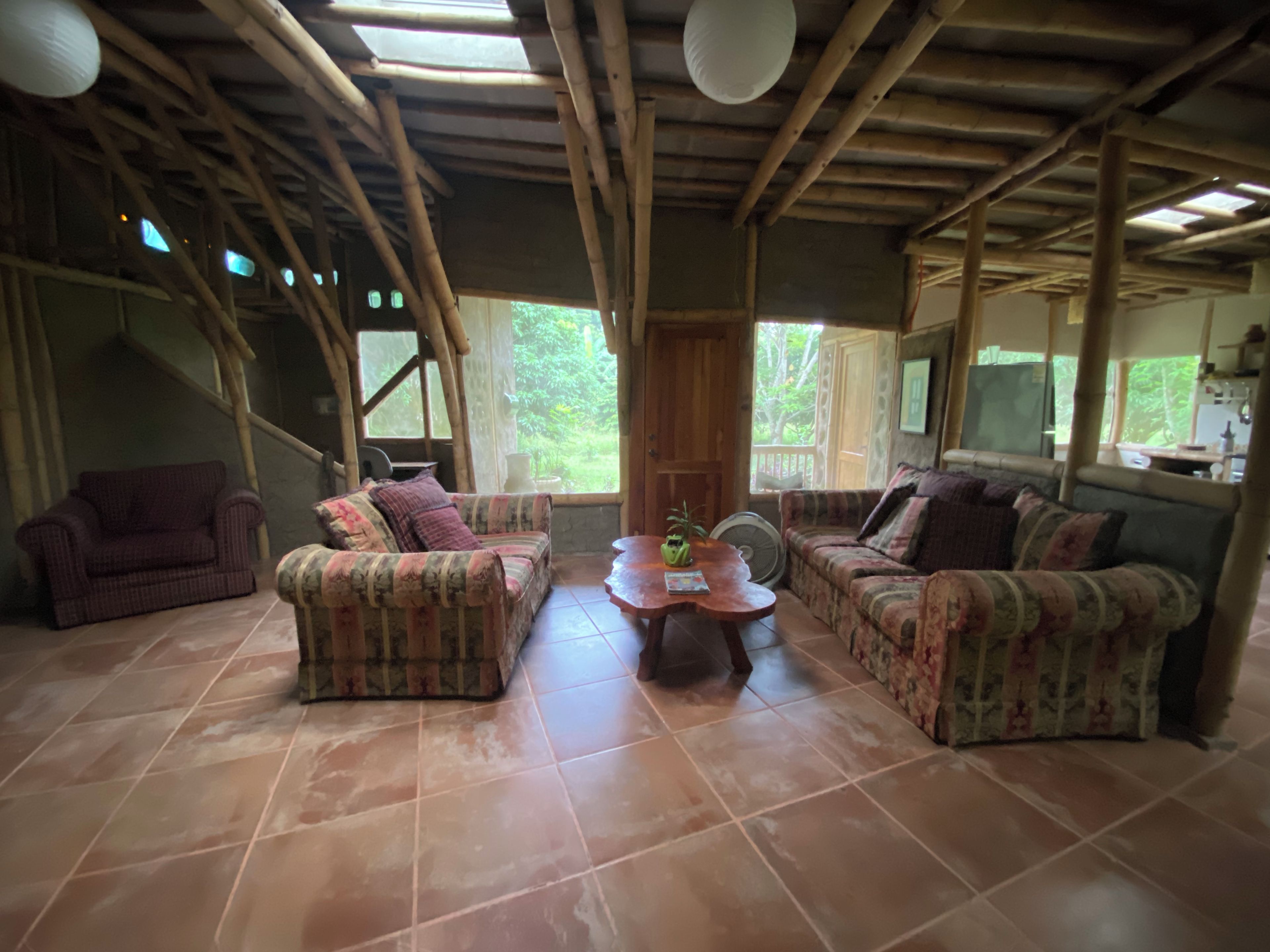 Quarto com teto alto, vigas de madeira e uma grande janela com vista para o exterior. Possui duas sofás estampadas e uma pequena mesa com um objeto decorativo.